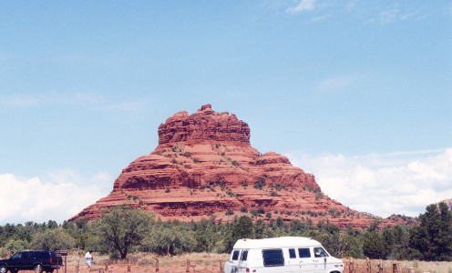 [A lone many-layered red rock rises from the ground in the shape of a bell. There are some green spots on it (growing vegetation), but it is prodominately red. There are trees all along the ground between the parking lot (where this image was taken) and Bell Rock. The top half of a white van in the parking lot is visible at the bottom edge of the photo. The sky is light blue with some white clouds at the level of the top of the trees and extends partway up the rock.]